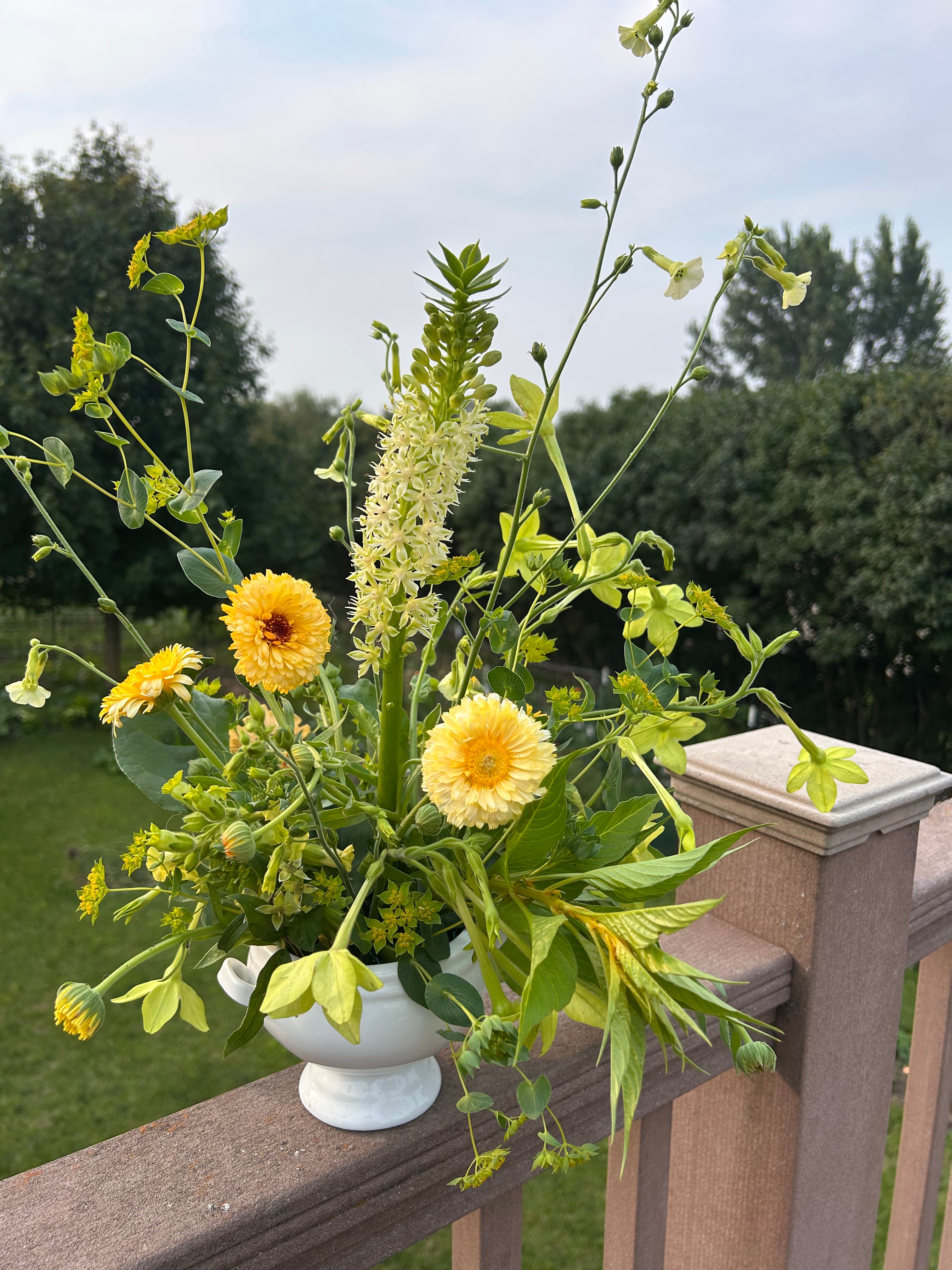 Bouquet of yellow flowers and green leaves in a white vase on a wooden deck.